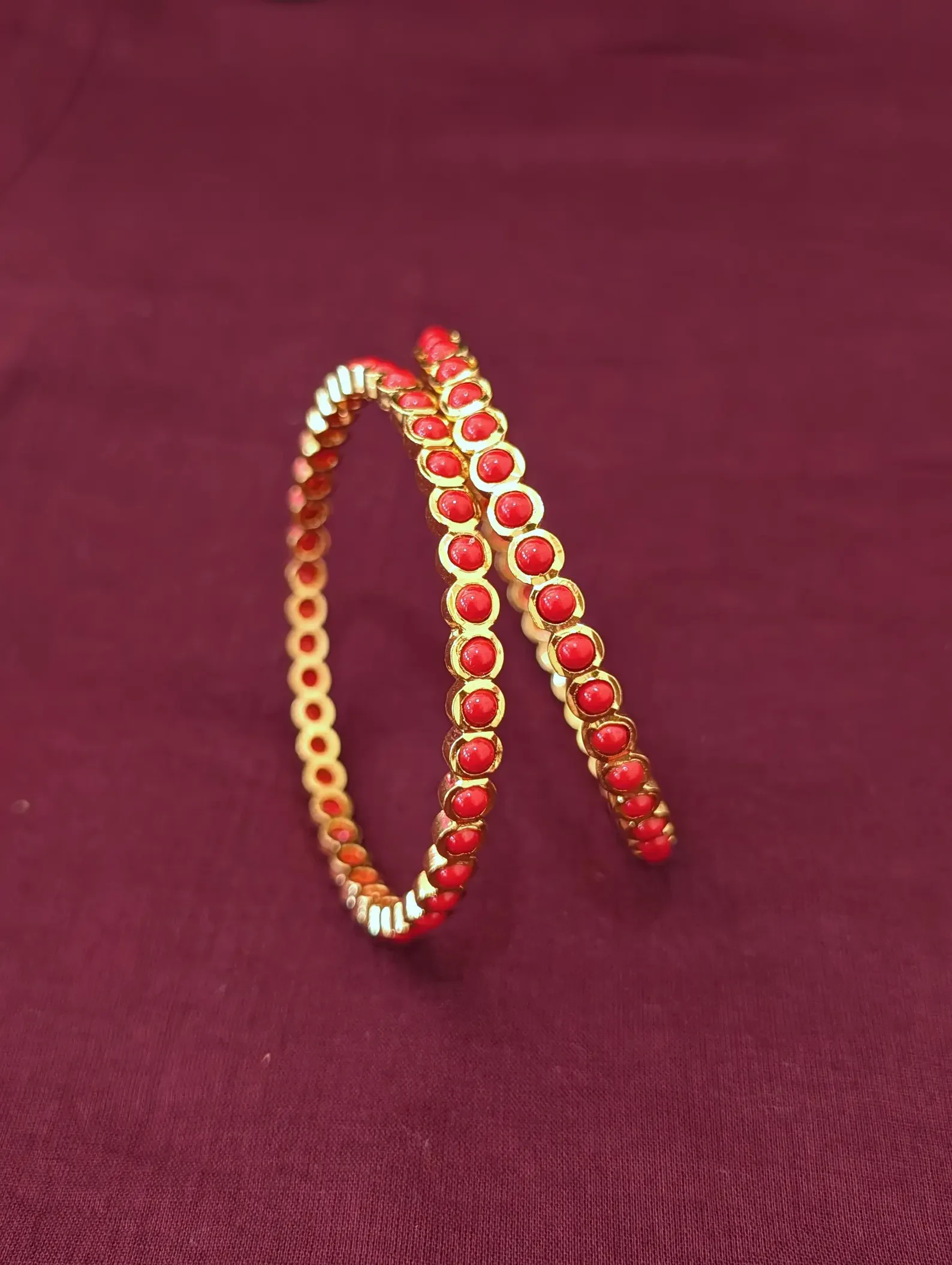 Pair of gold-tone bangles with coral-coloured round accents displayed upright on maroon fabric