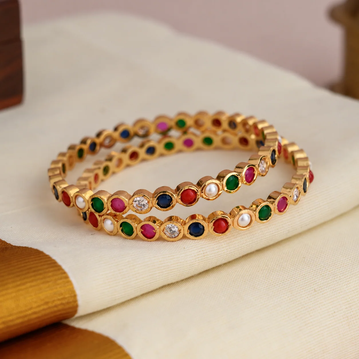 Pair of gold-tone multicolour stone bangles arranged on folded ivory fabric