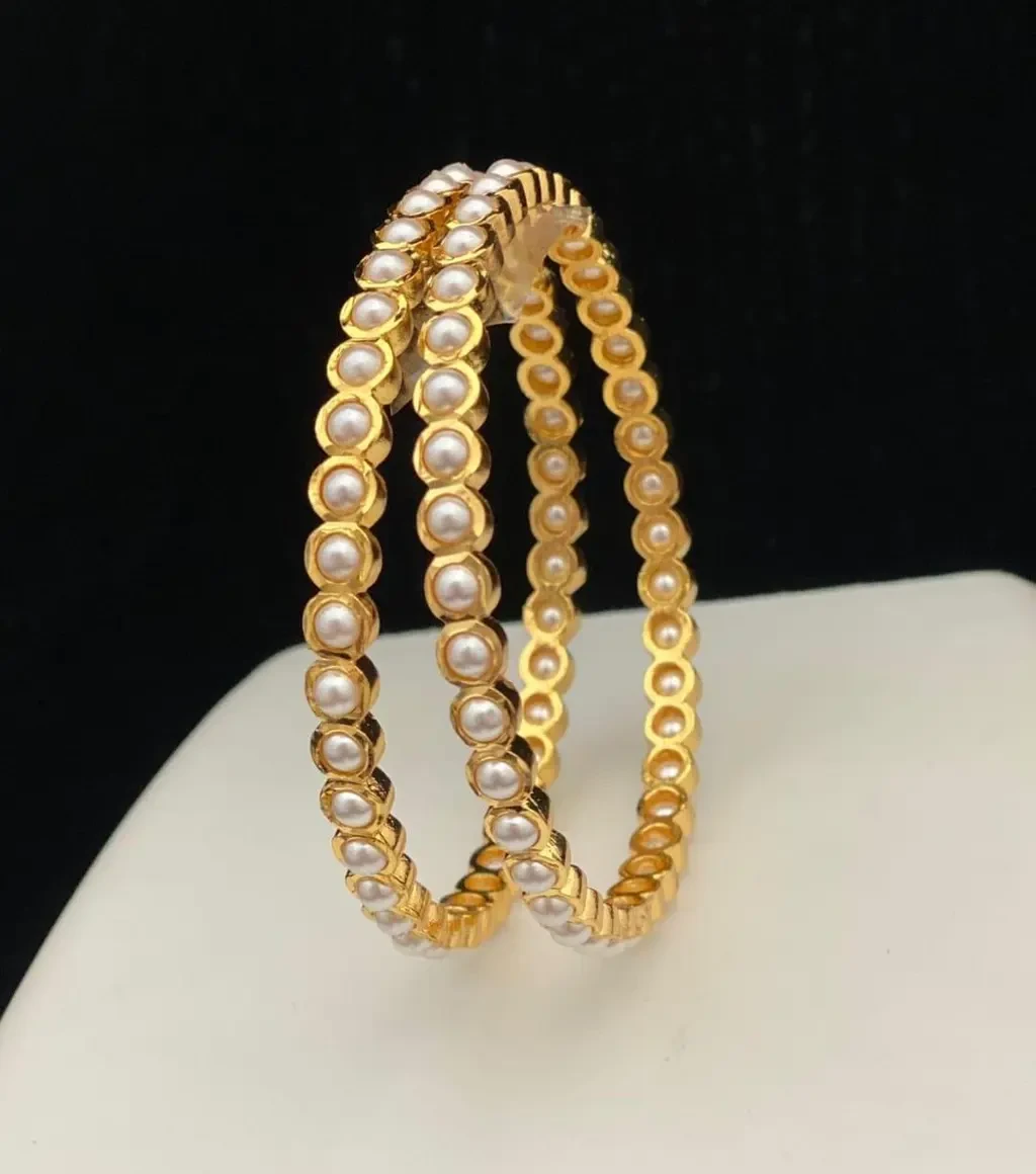 Pair of gold-tone bangles with round white pearl-like accents displayed upright against a dark background
