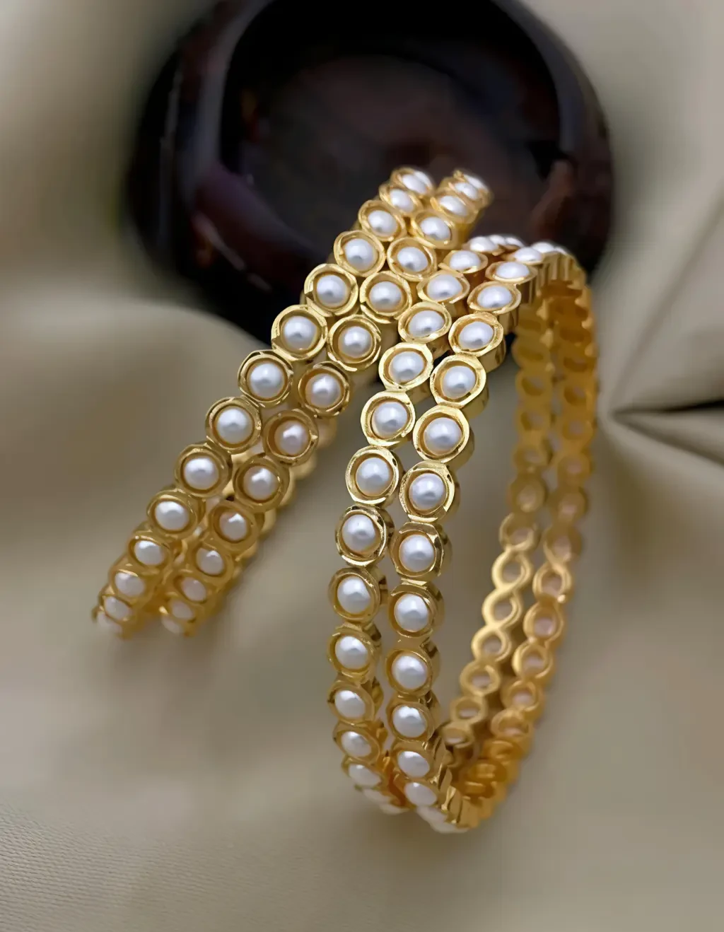 Pair of gold-tone bangles with round white pearl-like accents arranged on a pale cushioned surface